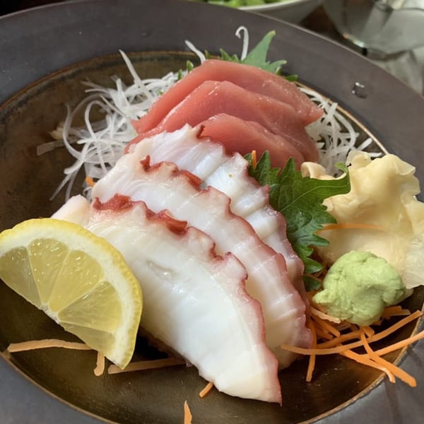 Sashimi platter with sliced tuna, octopus, lemon, ginger, and wasabi on a dark plate.