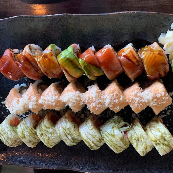 Three rows of colorful sushi rolls, including salmon, avocado, and seaweed-wrapped varieties, on a dark plate.